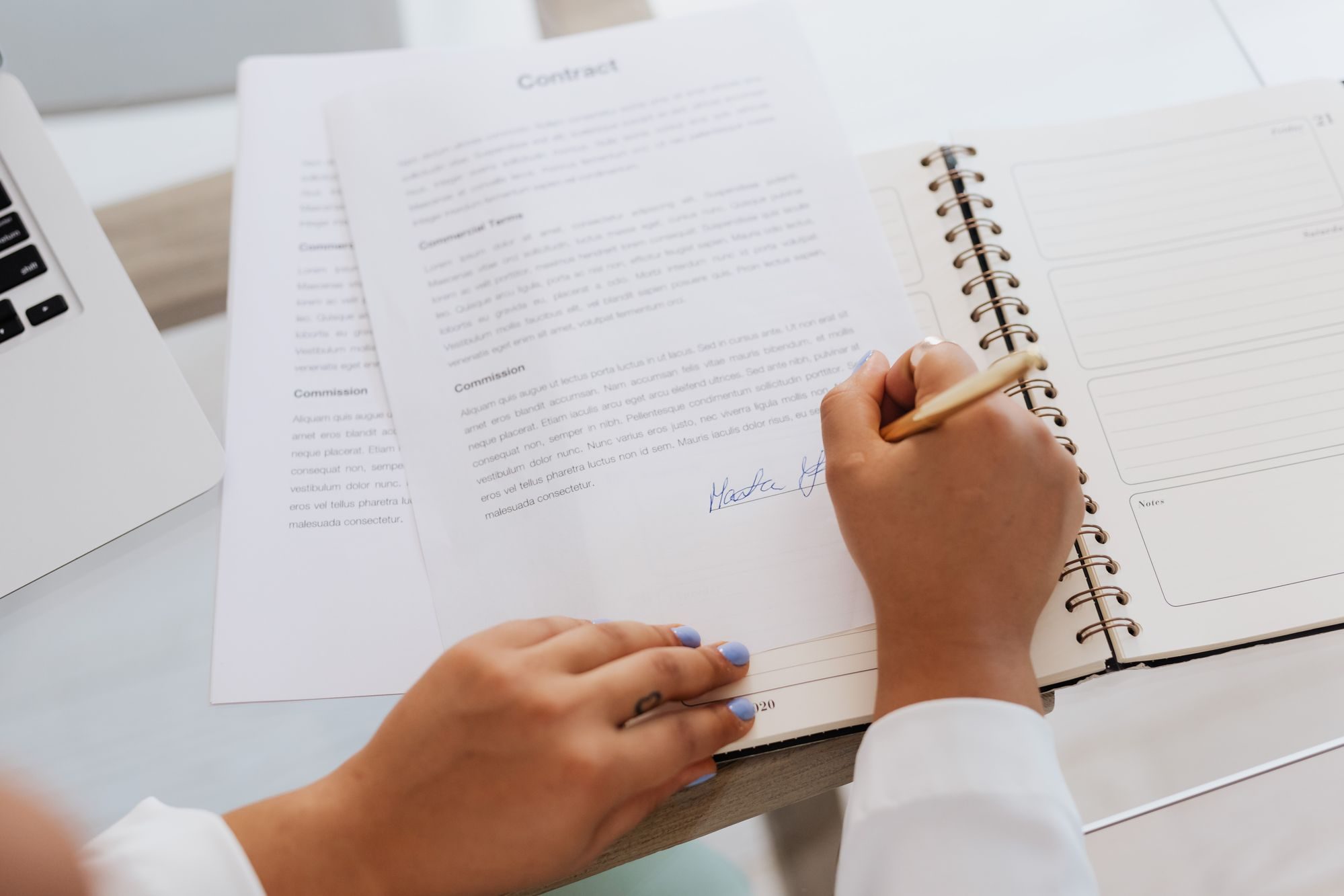 Dłonie podpisują dokument obok laptopa i notesu